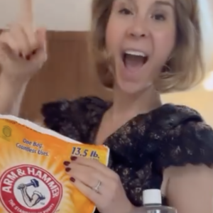 A woman excitedly points upward, holding a 13.5 lb bag of Arm & Hammer baking soda. She has short hair and is wearing a dark, textured dress.